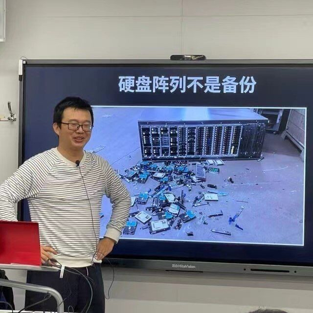续本达教授在 TUNA 活动中演讲，身后的屏幕上写着“硬盘阵列不是备份”，并展示倒地的硬盘柜和散落在地的硬盘
